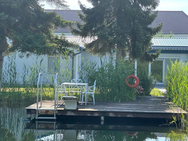 Gemütlicher Steg mit Tisch und Stühlen am ruhigen Gewässer, umgeben von Pflanzen.