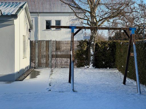 Schaukelgestell im Schnee neben einem weißen Haus und einem Zaun.