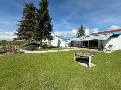 Landschaft mit einem gepflegten Garten, einem Wohnhaus und einem Grillplatz unter blauen Himmel.