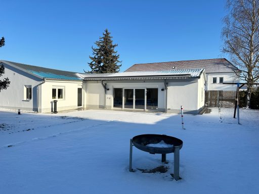 Helles Haus mit Terrasse und Grill im Schnee, umgeben von Bäumen.