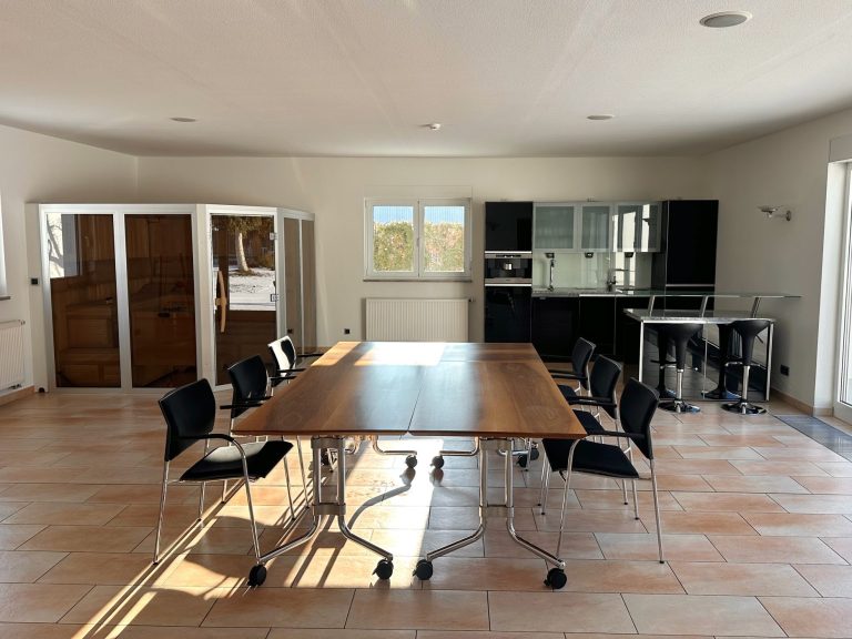 Großer, heller Raum mit einem langen Tisch und Stühlen, angrenzender Küche.