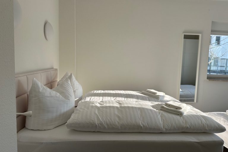 Helles Schlafzimmer mit einem Doppelbett, weißen Kissen und einem Handtuch auf der Bettdecke.