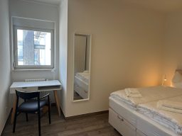 Helles Zimmer mit Doppelbett, Tisch und Stuhl sowie einem Spiegel und Fenster.