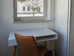 Weißer Schreibtisch mit Holzbeinen und einem braunen Stuhl, Fenster im Hintergrund.